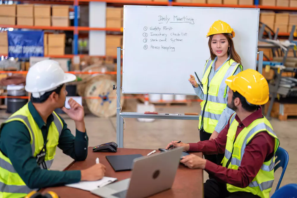 A woman wearing a safety helmet and high-visibility vest giving a presentation at a whiteboard labelled "Daily Meeting," while two colleagues in similar safety gear listen attentively at a table in a warehouse setting.