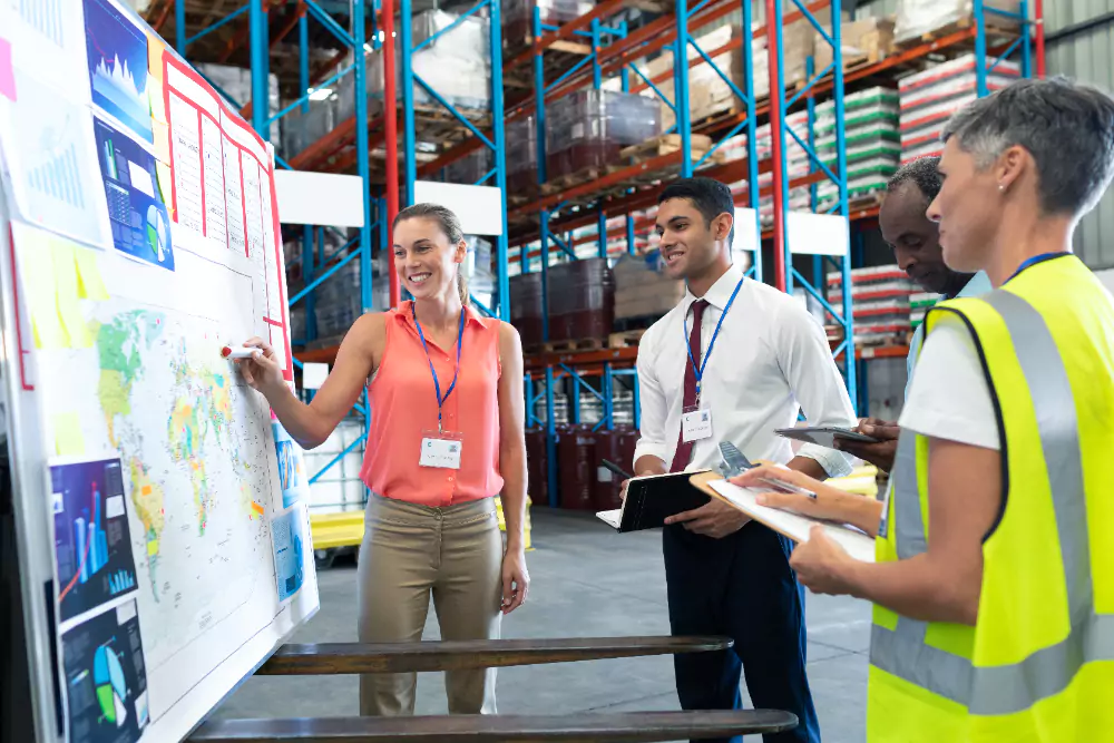 A group of professionals in a warehouse setting, gathered around a whiteboard with charts, maps, and graphs. A woman wearing a coral-coloured blouse and a name badge is leading the discussion, pointing at a map with a red marker.
