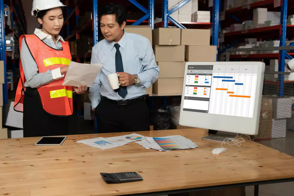 A man in formal attire and a woman wearing a safety vest and helmet review documents together in a warehouse. A desk nearby holds a computer displaying a project timeline, along with charts, a tablet, and a calculator. Shelves filled with boxes are visible in the background.