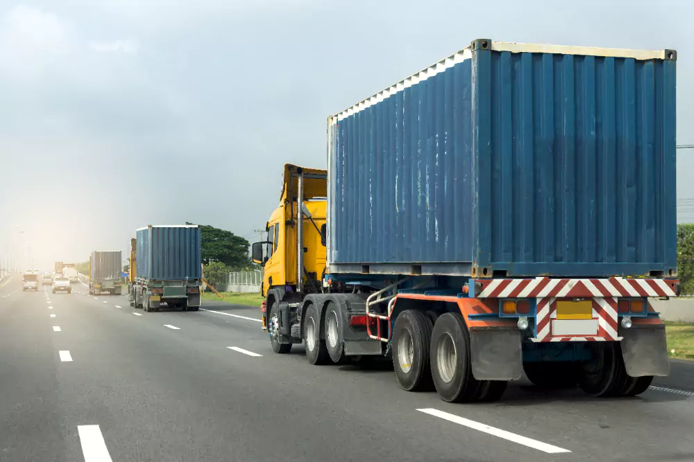Lorries carrying blue shipping containers travelling on a multi-lane highway, with a cloudy sky in the background.
