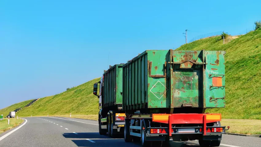 A green freight truck carrying a large container on a highway, surrounded by lush green hills under a clear blue sky. Ideal depiction of road freight transport in the UK.